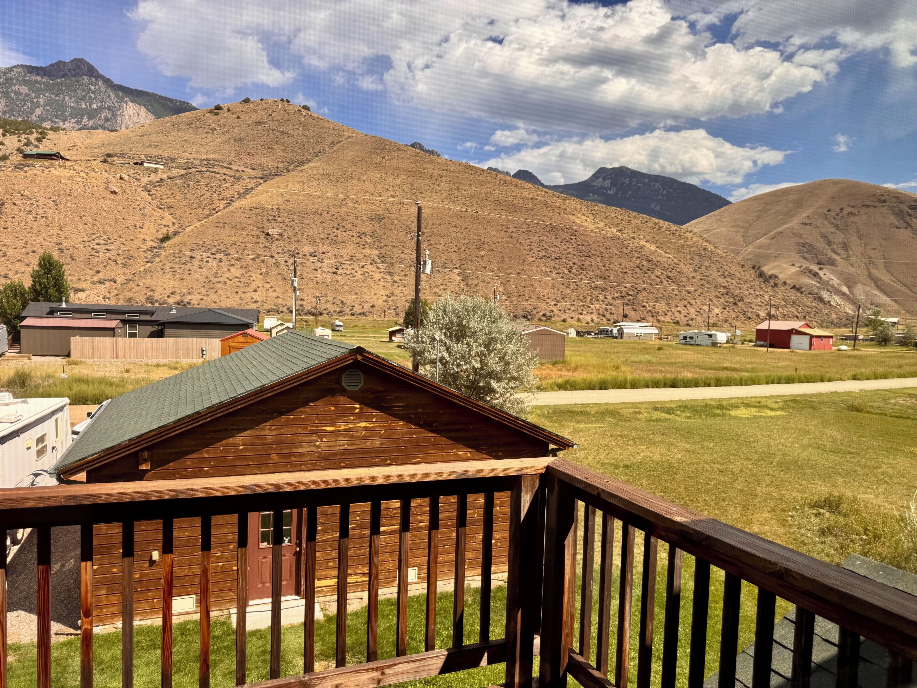 A wooden cabin with a fenced deck, surrounded by a grassy field and mountains in the background.