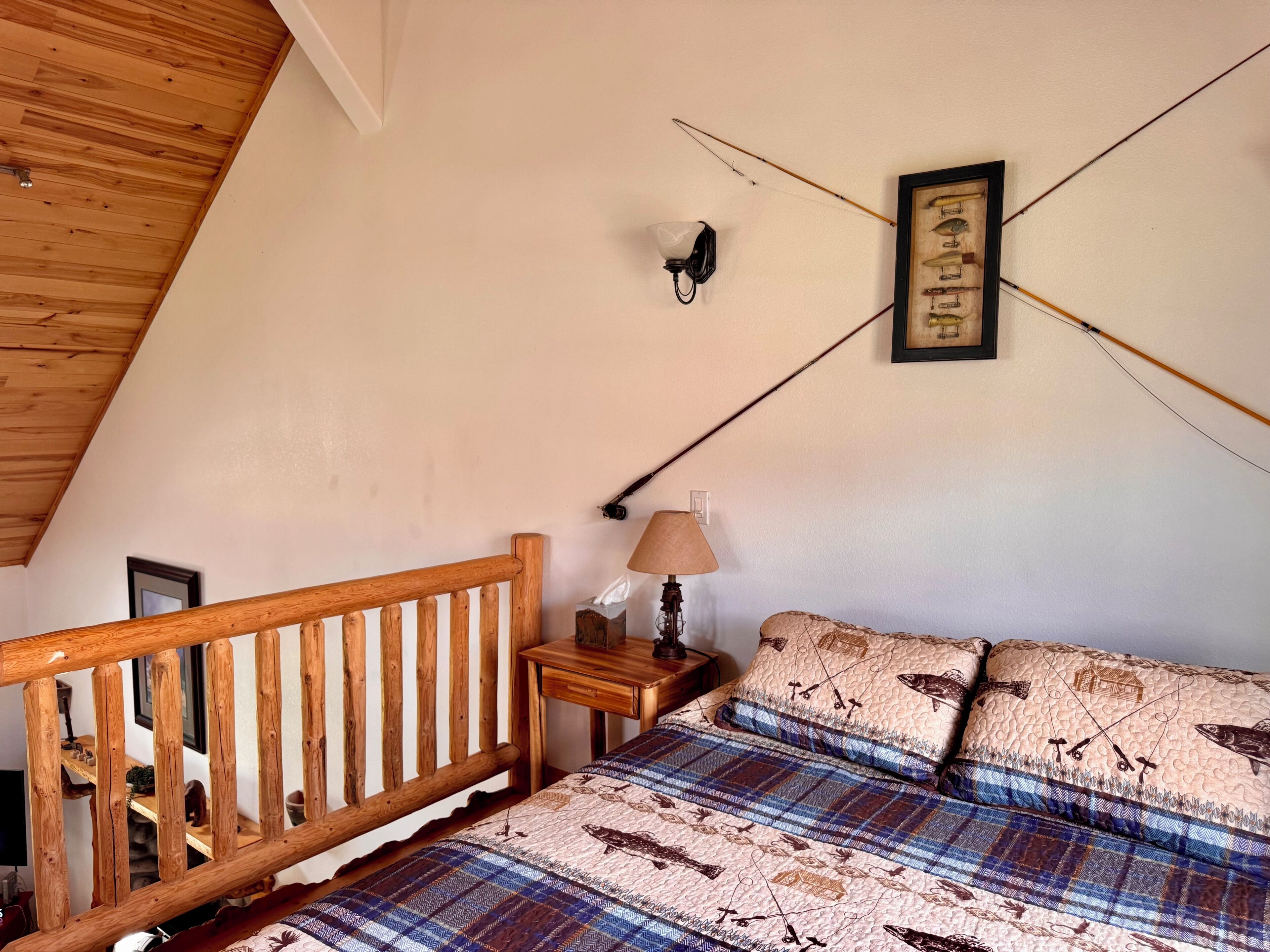 Quarto com teto de madeira, cama com colcha xadrez, mesa de cabeceira com abajur e luminária fixada na parede.