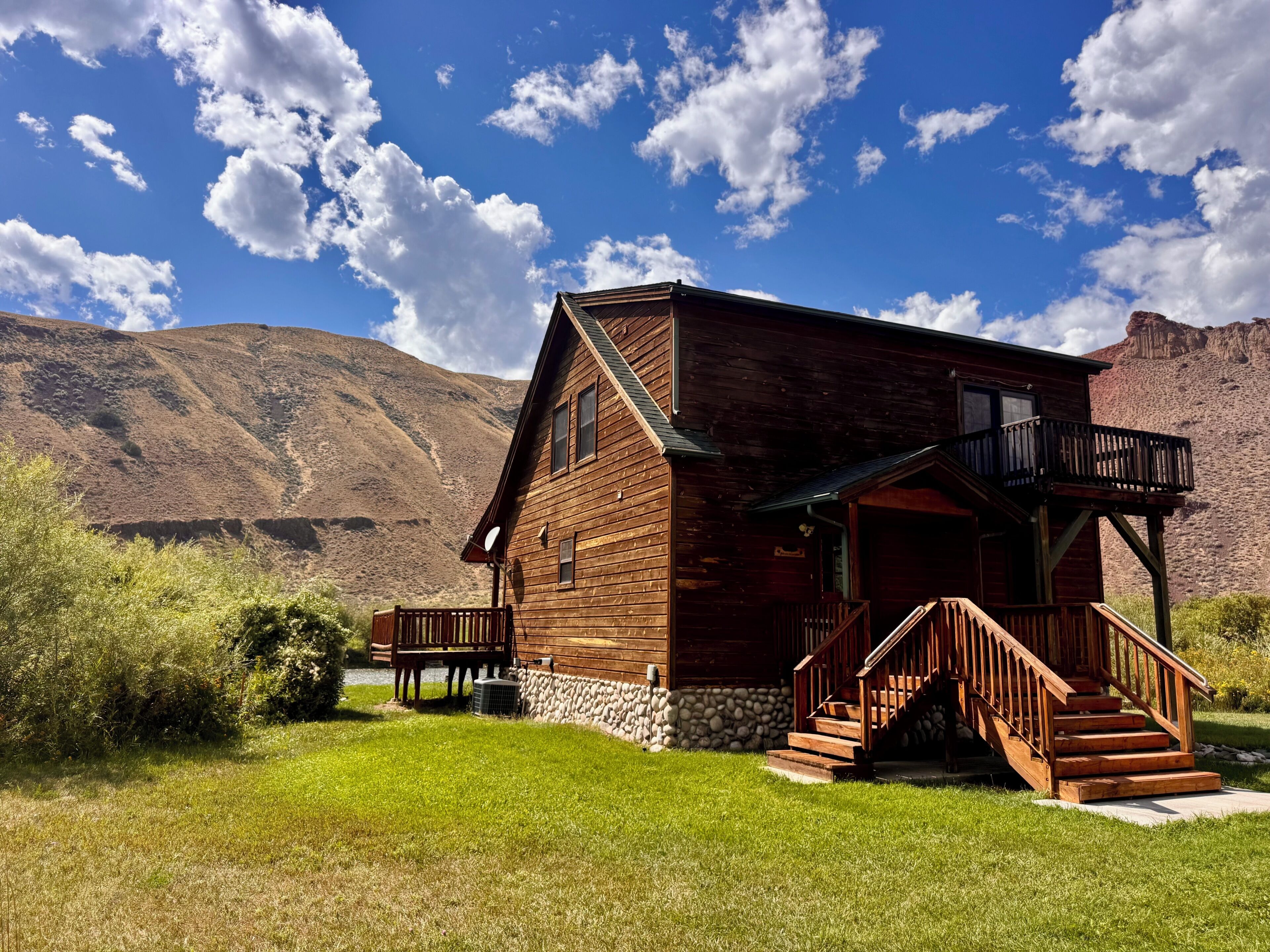 A wooden cabin with a porch and stairs, surrounded by a grassy area and mountains in the background.