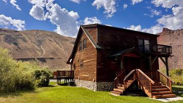 A wooden cabin with a porch and stairs, surrounded by a grassy area and mountains in the background.