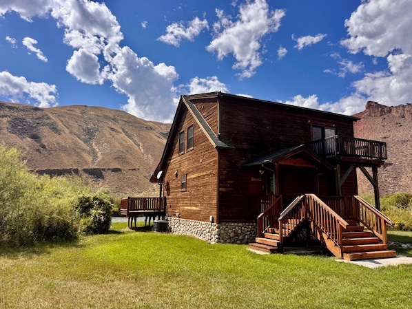 A wooden cabin with a porch and stairs, surrounded by a grassy area and mountains in the background.