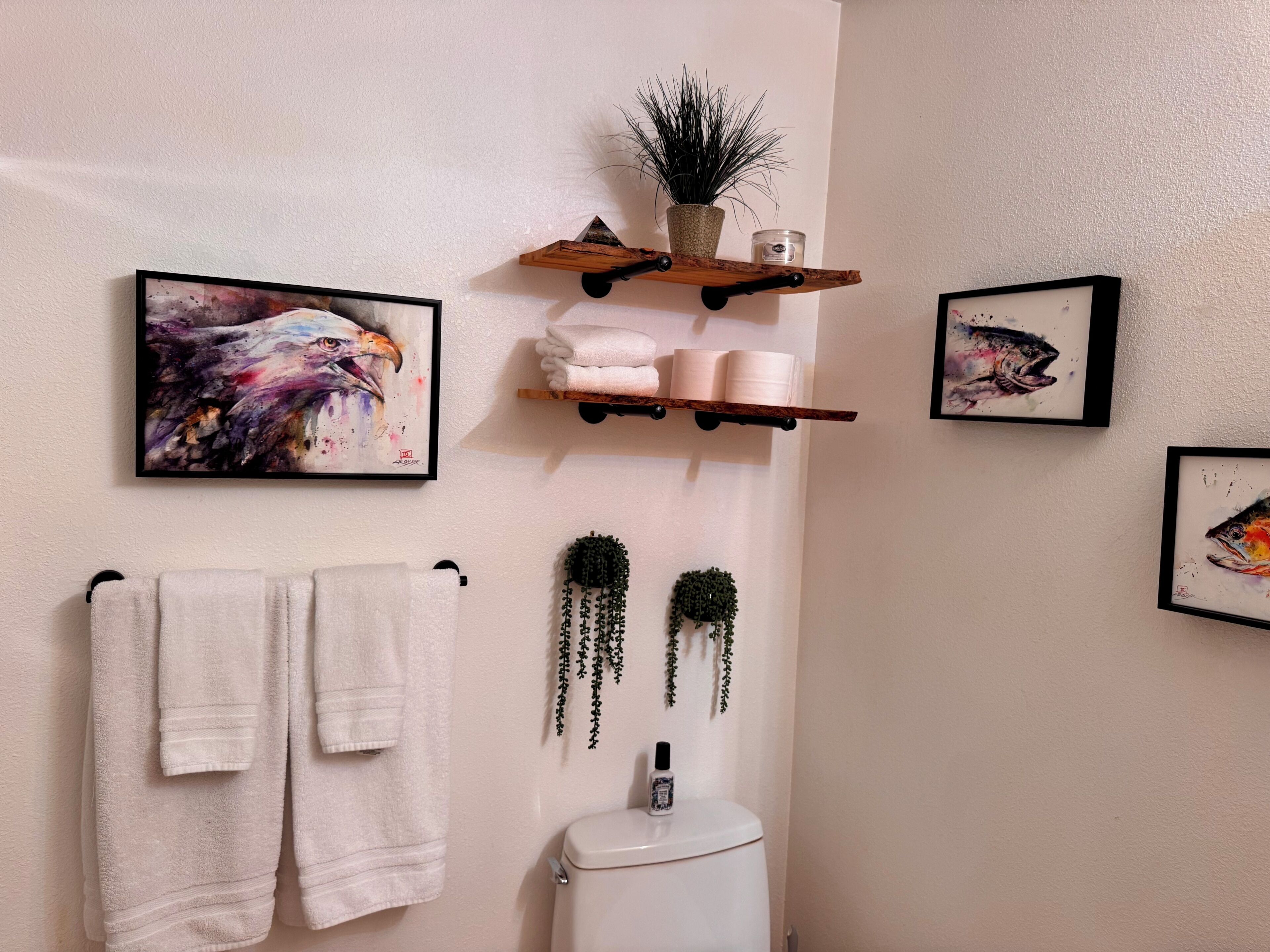A bathroom with a toilet, two towels, a shelf with toiletries, and three framed artworks.