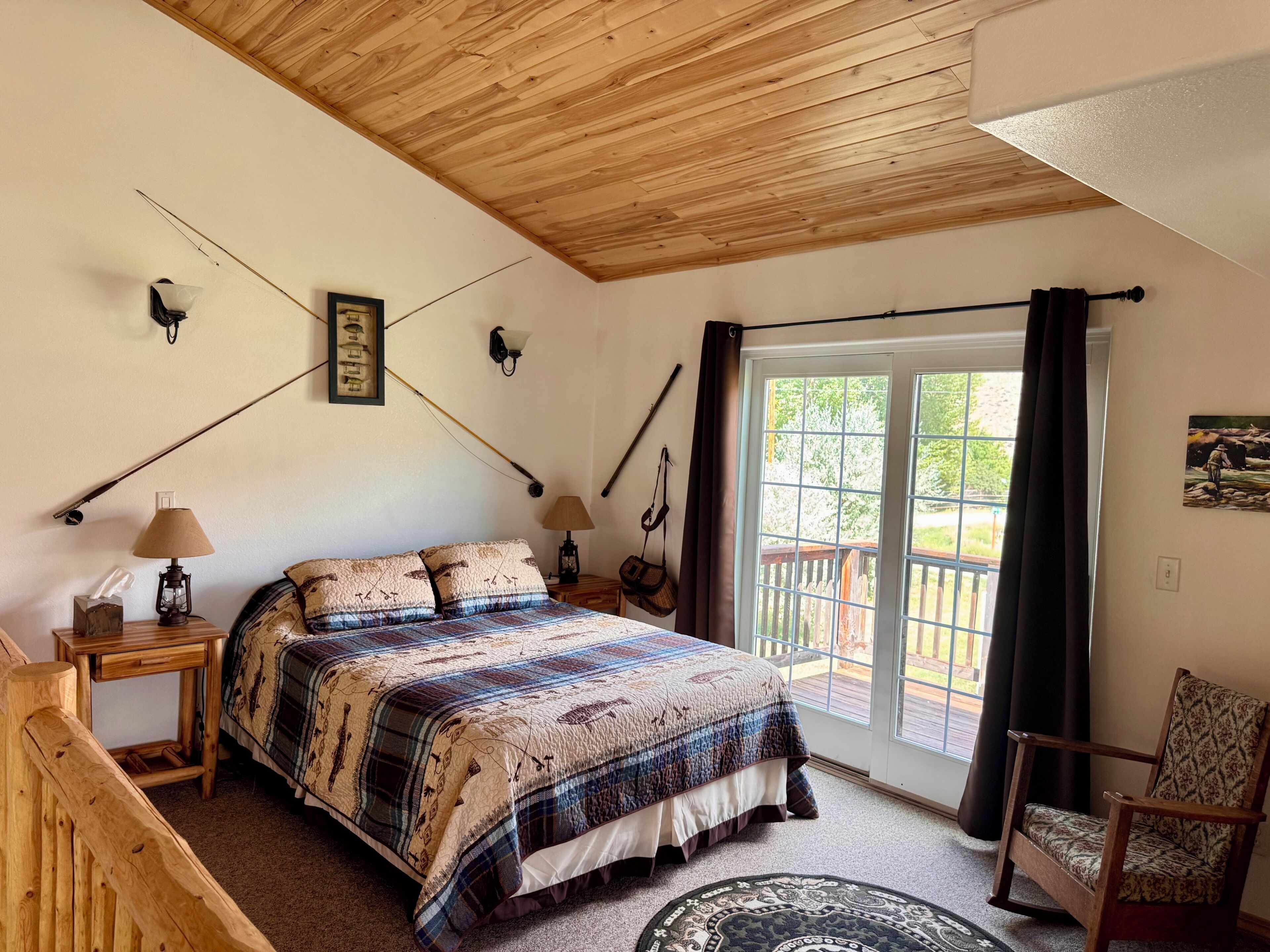A bedroom with a wooden ceiling, a bed with a patterned bedspread, a nightstand with a lamp, and a chair.