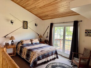 A bedroom with a wooden ceiling, a bed with a patterned bedspread, a nightstand with a lamp, and a chair.
