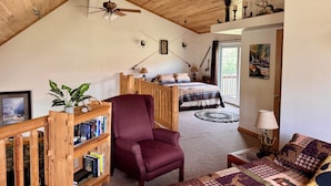 Habitació acollidora amb sostre inclinat, una prestatgeria de fusta, un sofà còmode, un llit amb un estriatge i un quadre a la paret.