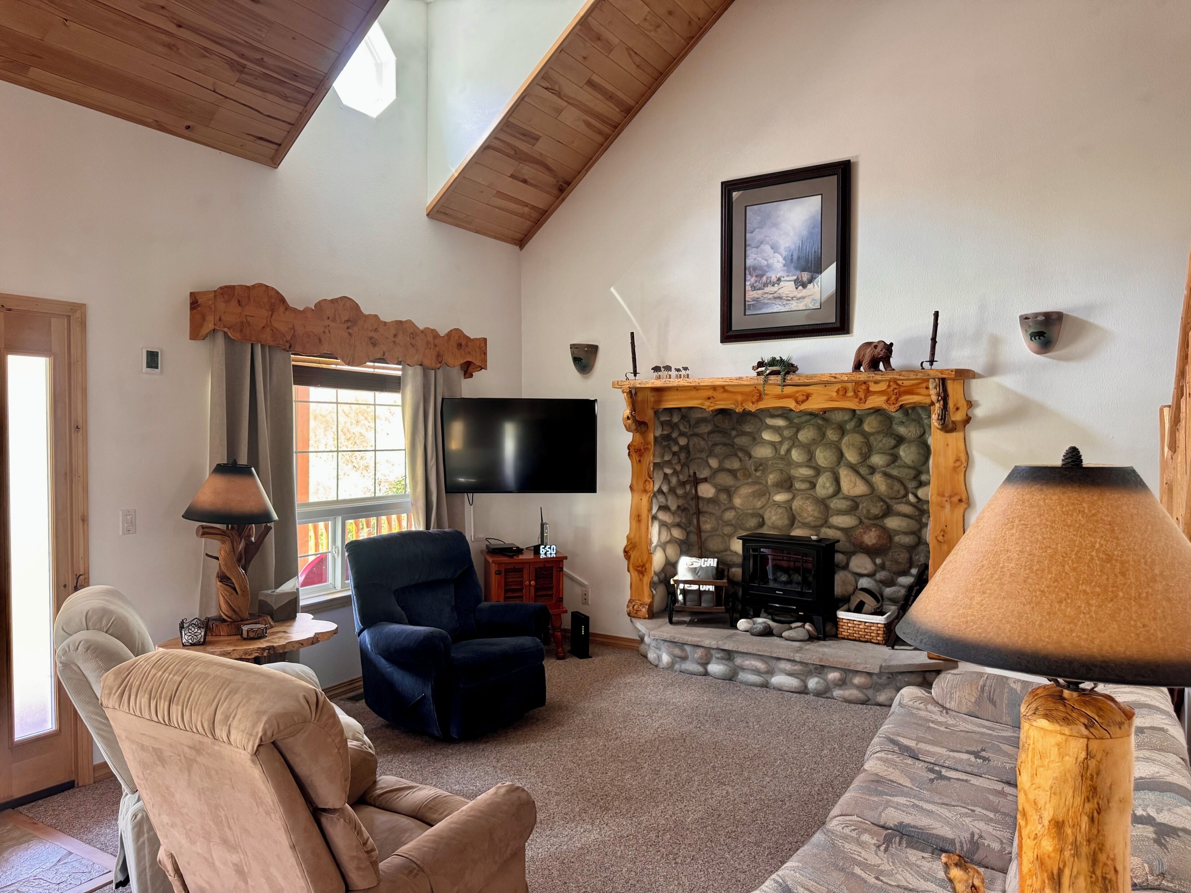 A living room with a stone fireplace, a television mounted on the wall, and a large window with curtains.