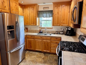 A kitchen with wooden cabinets, a stainless steel refrigerator, a sink, a stove, and a window with a view of greenery.