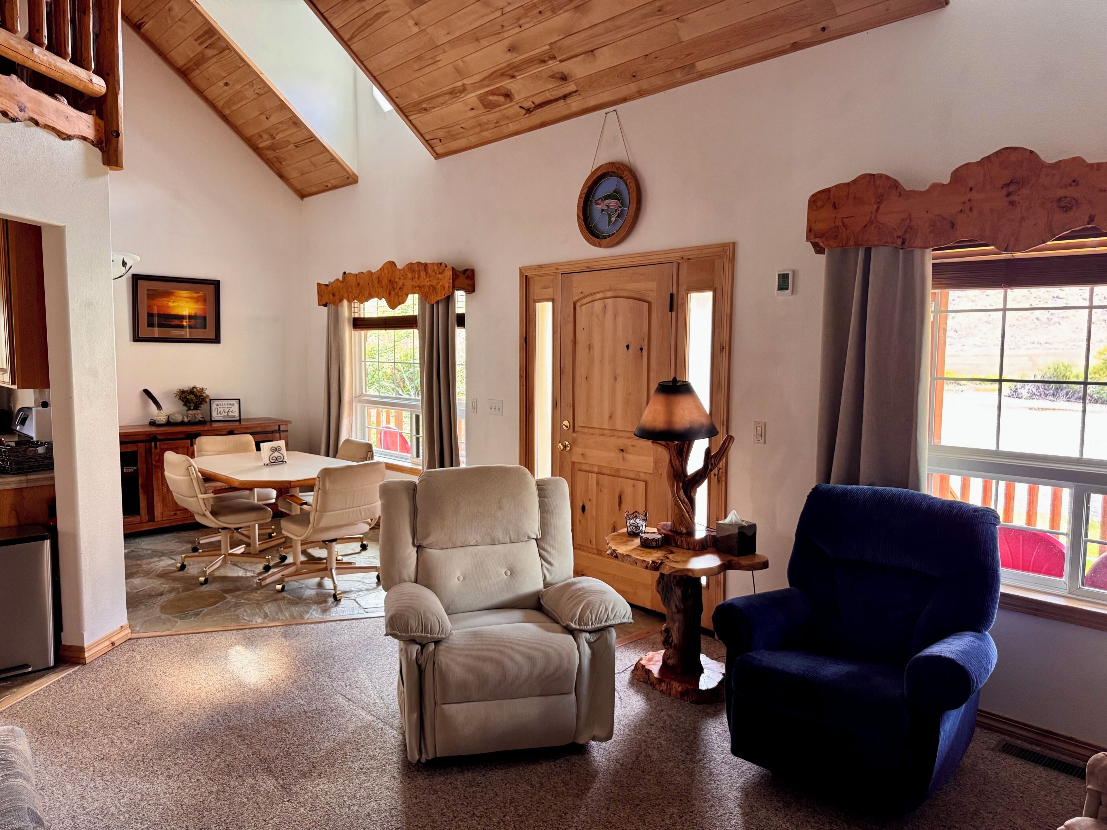 A living room with a dining area, a wooden ceiling, a clock, and a window with curtains.
