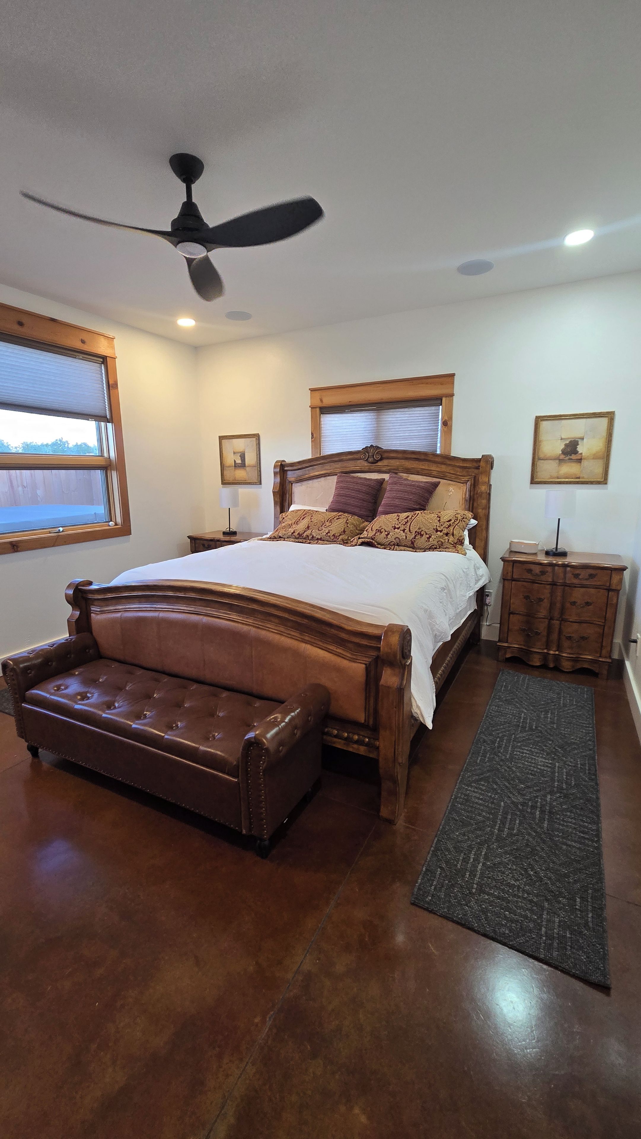 A bedroom with a large bed, a ceiling fan, a nightstand, a dresser, and a window with blinds.