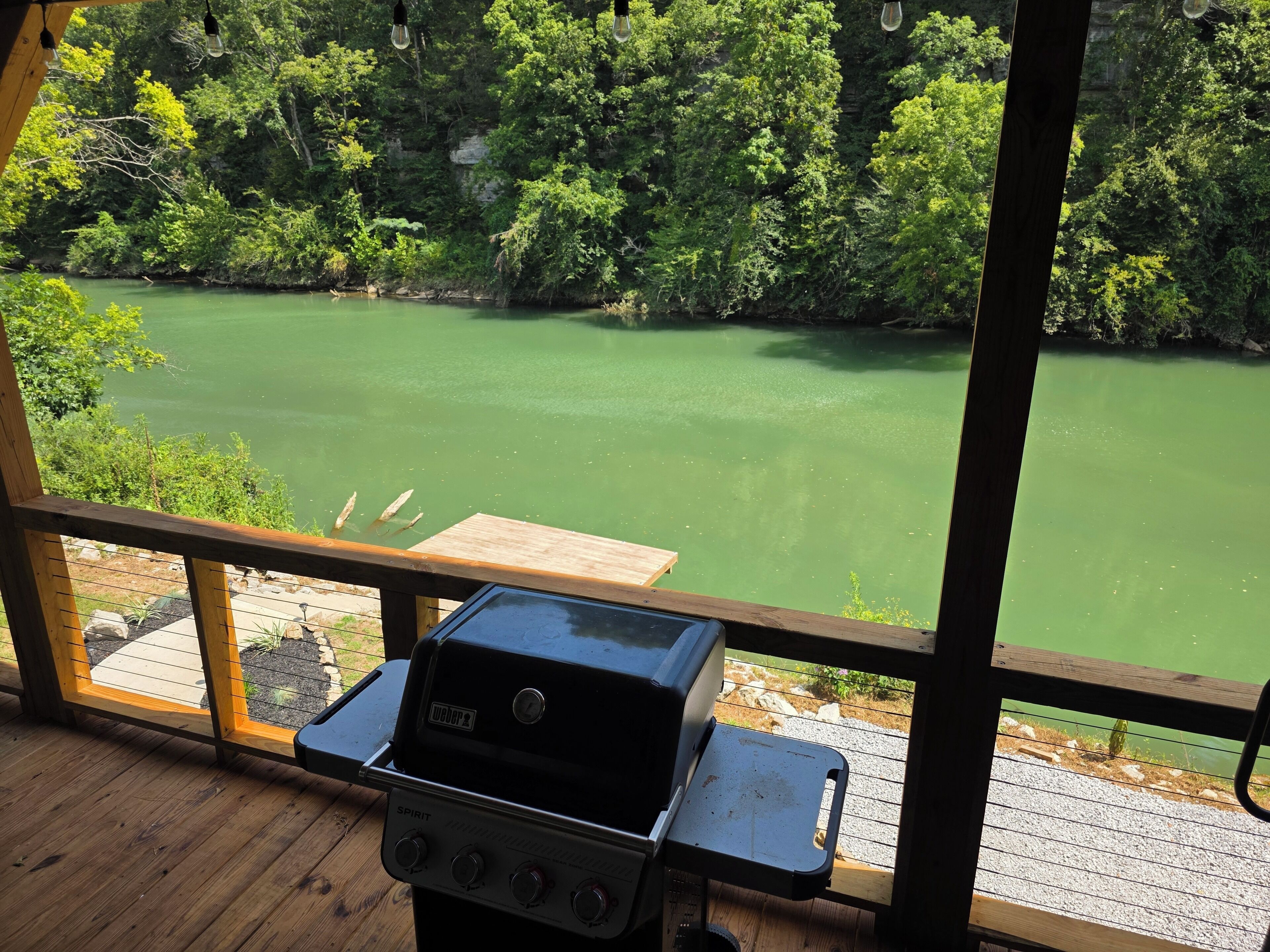 A wooden deck with a grill and a view of a green lake surrounded by trees.