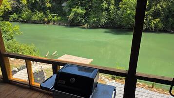 A wooden deck with a grill and a view of a green lake surrounded by trees.