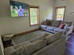 A living room with a large flat-screen TV mounted on the wall, a grey sofa, a patterned rug, and a window with wooden frames. - Charming 2-bedroom cabin on the water with AC in Linden (Linden)