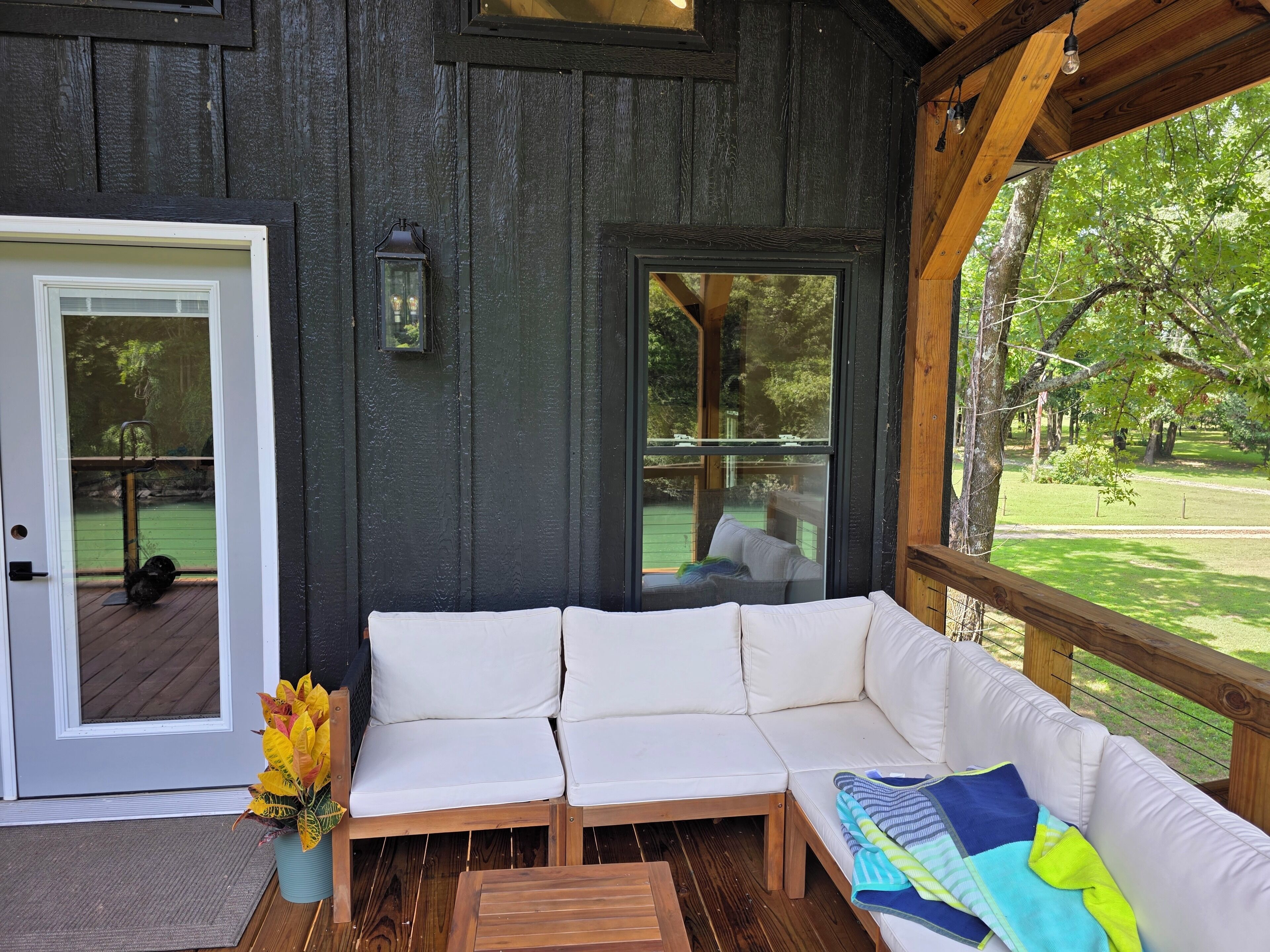 Eine Holzterrasse mit einer weißen Couch und einem kleinen Tisch, eine Tür mit Blick auf eine grüne Rasenfläche und ein großes Fenster mit Blick auf Bäume.