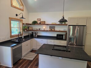 A kitchen with white cabinets, a stainless steel refrigerator, a black countertop, and wooden shelves with various items. - Charming 2-bedroom cabin on the water with AC in Linden (Linden)