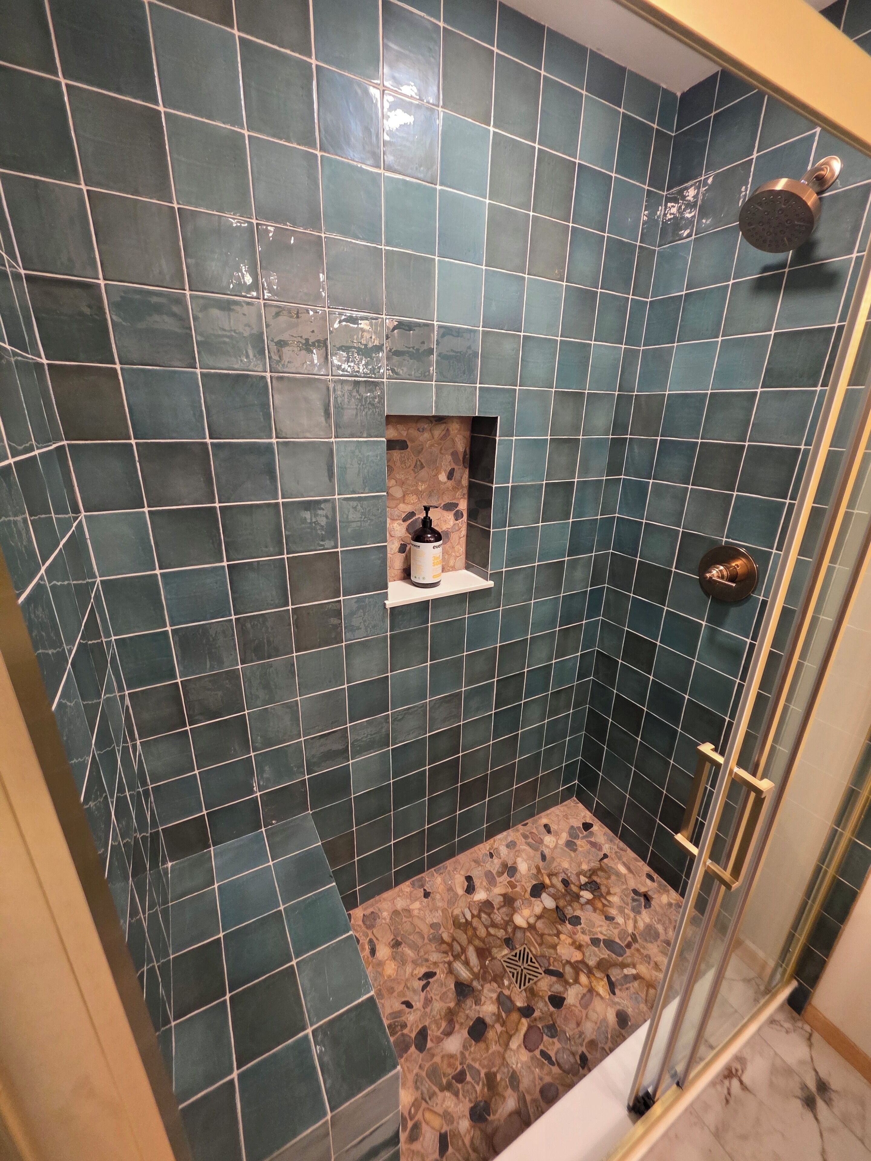 Una ducha con azulejos azul oscuro, un estante pequeño con una botella y una puerta de vidrio.