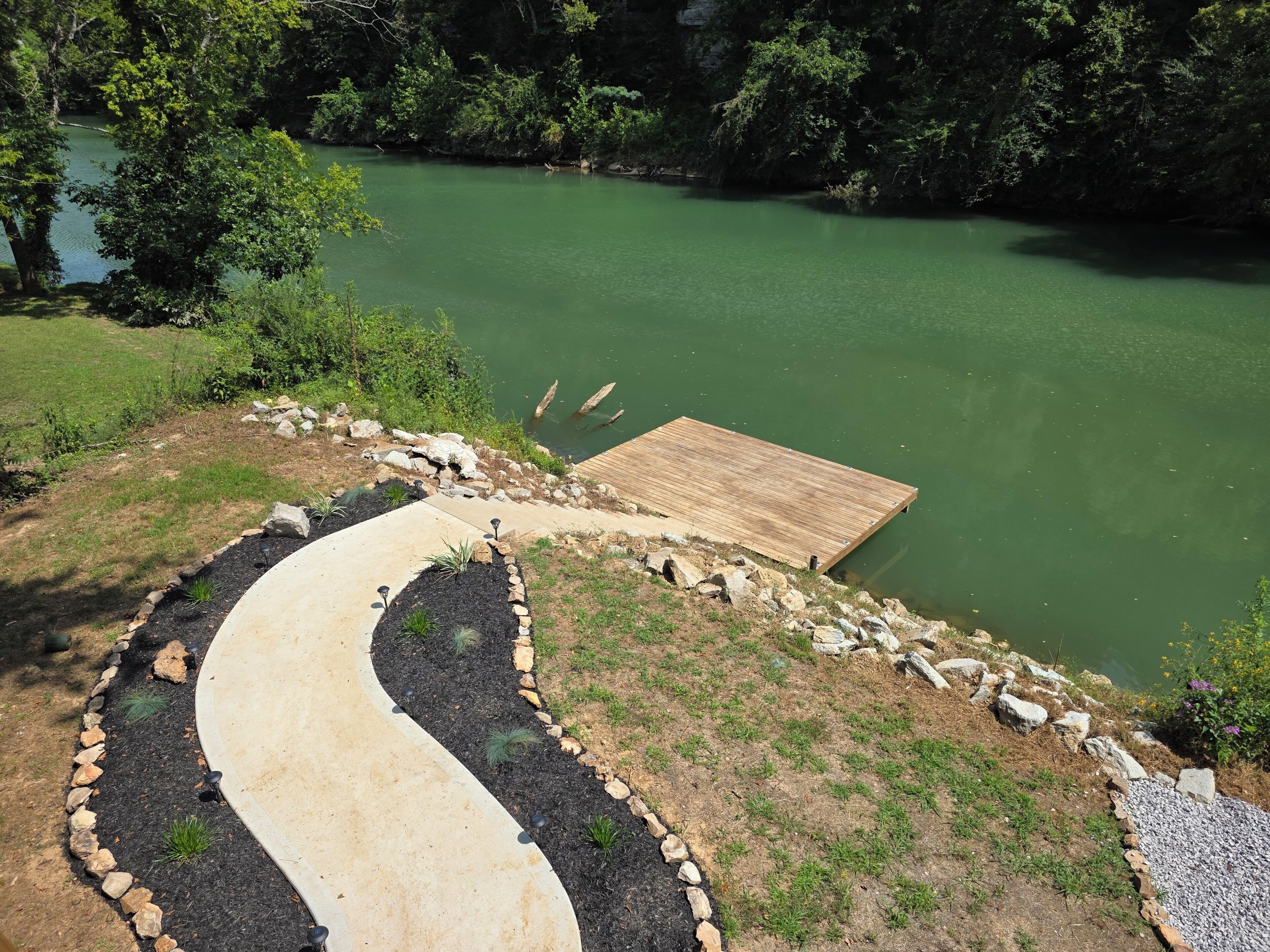A wooden dock extending into a green lake, surrounded by a landscaped area with a winding path and stone borders.