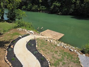 A wooden dock extending into a green lake, surrounded by a landscaped area with a winding path and stone borders.