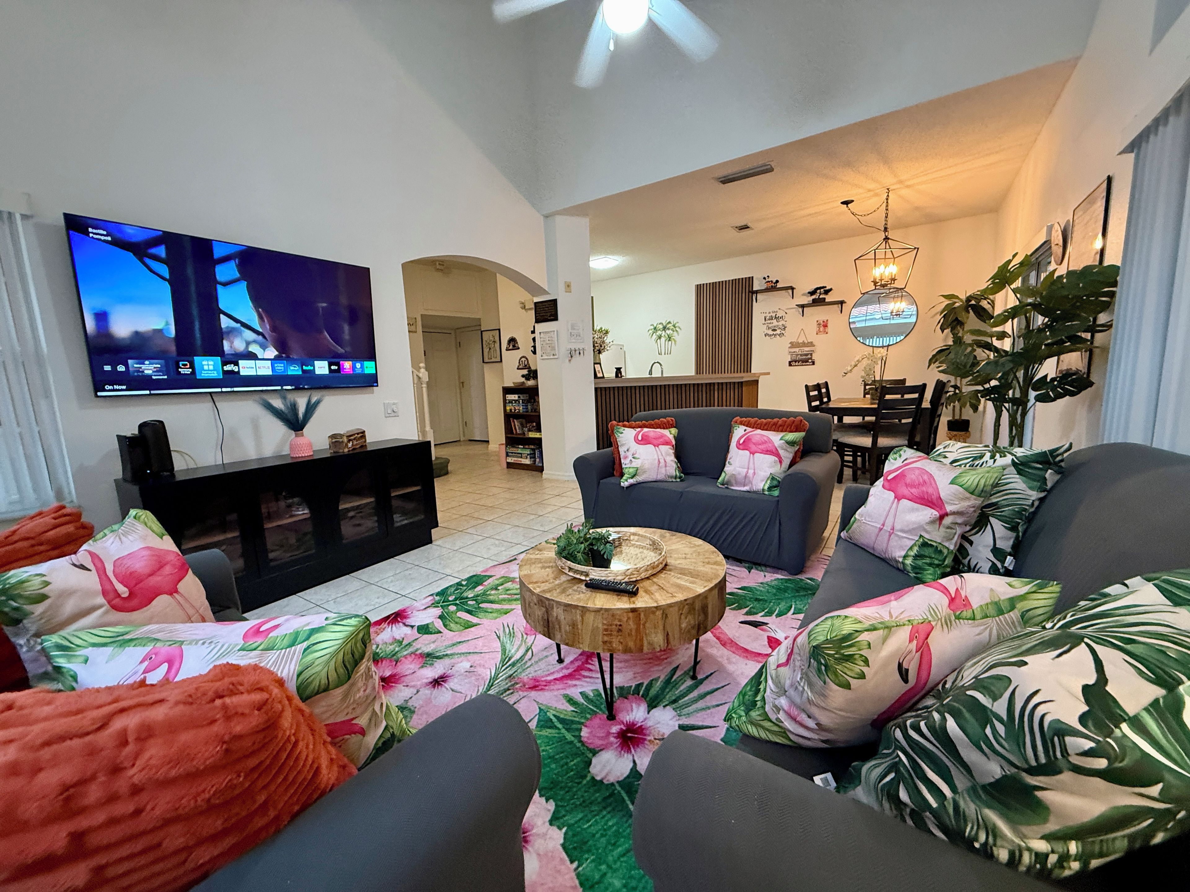 A living room with a flat-screen TV, a sofa set with floral cushions, a wooden coffee table, and a dining area in the background.