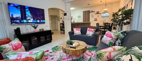 A living room with a flat-screen TV, a sofa set with floral cushions, a wooden coffee table, and a dining area in the background.
