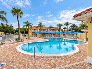 A swimming pool with lounge chairs and umbrellas, surrounded by palm trees and buildings with red-tiled roofs. - 20 Min to Disney World | 4BR Pool Home (Kissimmee)