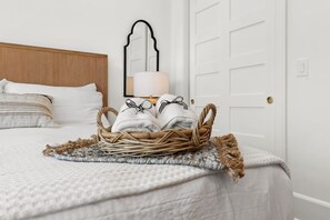 A neatly made bed with a white comforter and pillows, a wicker basket with folded towels, and a lamp on a bedside table. - This Must Be The Place (Ocean Isle Beach)
