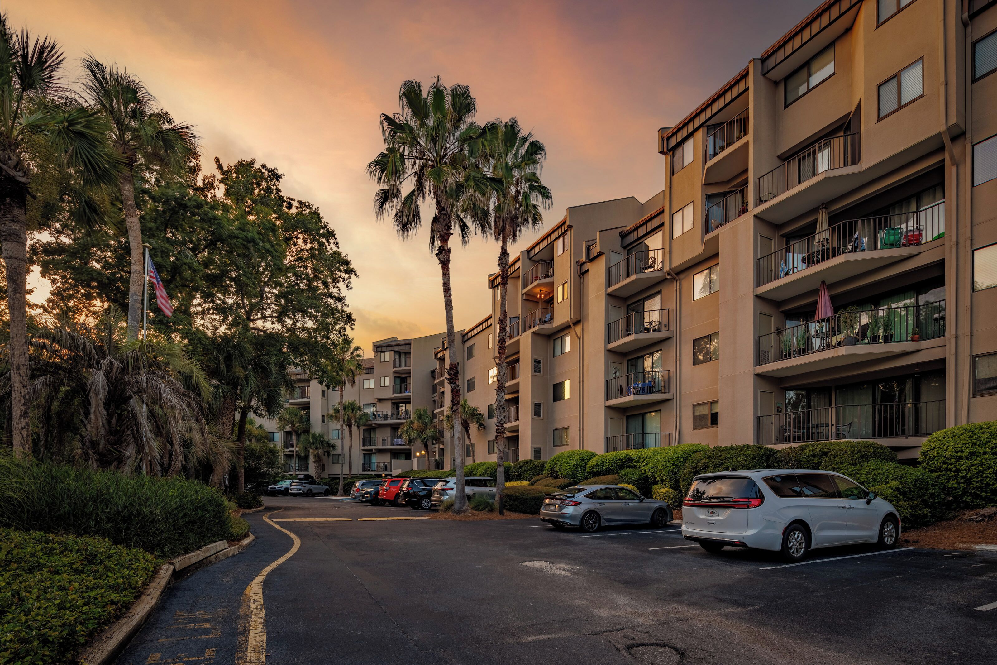 A multi-story residential building with balconies, a parking area, and palm trees.