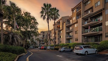 A multi-story residential building with balconies, a parking area, and palm trees.