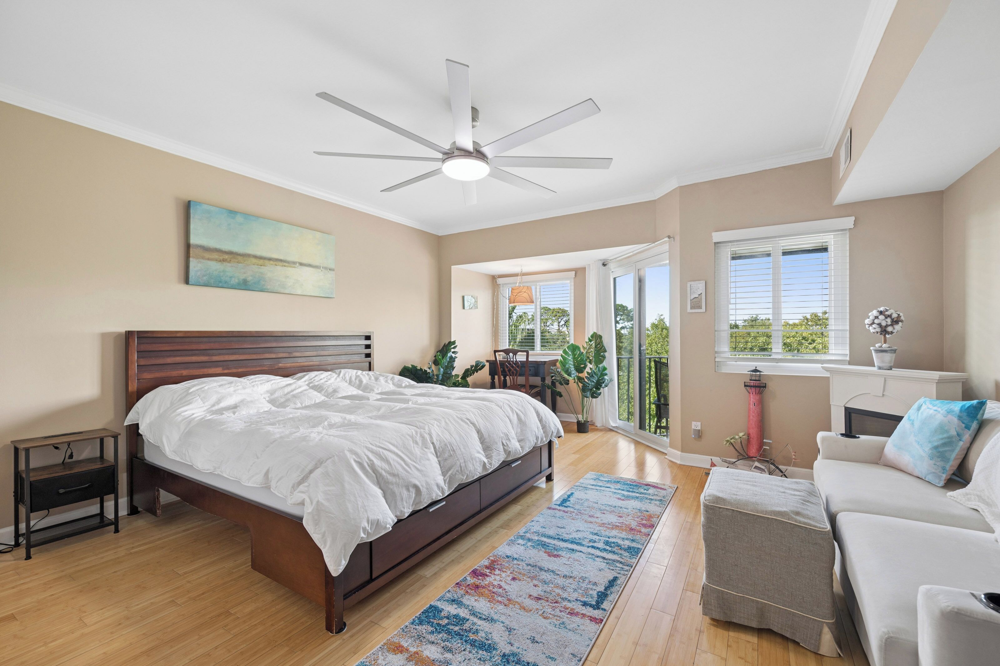 A bedroom with a large bed, a ceiling fan, a painting, a desk with a chair, and a view of the outdoors through a sliding door.