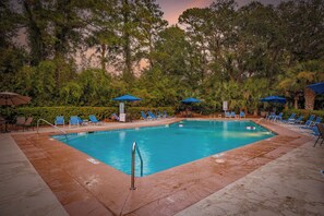 Piscina retangular com água azul, rodeada por espreguiçadeiras e guarda-sóis, com árvores e céu ao pôr do sol ao fundo.