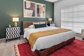 A bedroom with a bed, two bedside tables, a red rug, and a window with blinds.