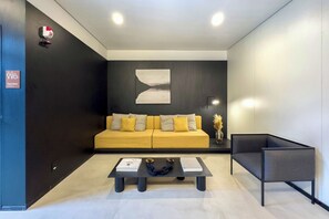 Un espace d’attente moderne avec un canapé jaune, une table basse avec des livres et une corde, un fauteuil noir et un luminaire fixé au mur.