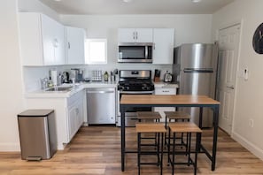 A modern kitchen with white cabinets, stainless steel appliances, a wooden dining table, and a trash bin. - Cozy 2-bedroom house in tranquil Clovis with AC (Clovis)