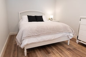 A white bed with a textured blanket, two black pillows, a bedside lamp, and a wooden floor. - Cozy 2-bedroom house in tranquil Clovis with AC (Clovis)
