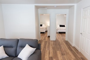 A modern interior with a grey sofa, wooden flooring, and two bedrooms with white walls and beds.