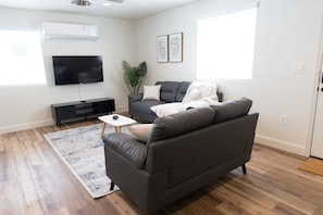 A modern living room with a grey sofa, a flat-screen TV, a small white side table, and a large window. - Cozy 2-bedroom house in tranquil Clovis with AC (Clovis)