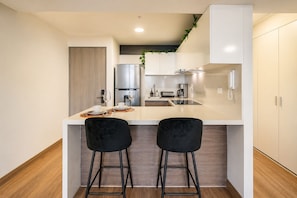 A compact kitchen with a white countertop, two black bar stools, a stainless steel refrigerator, a coffee maker, and a telephone. - Suite ejecutiva Piscina y Gym San Isidro (San Isidro)