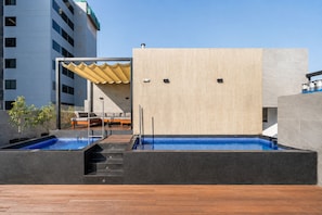 A modern outdoor pool area with a wooden deck, a seating area with a sofa and chairs, and a building with a large wooden panel wall. - Suite ejecutiva Piscina y Gym San Isidro (San Isidro)