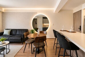 Un salon moderne avec un canapé en cuir noir, une table de salle à manger ronde entourée de chaises, et un coin cuisine doté d’un plan de travail et de meubles de rangement en bois.