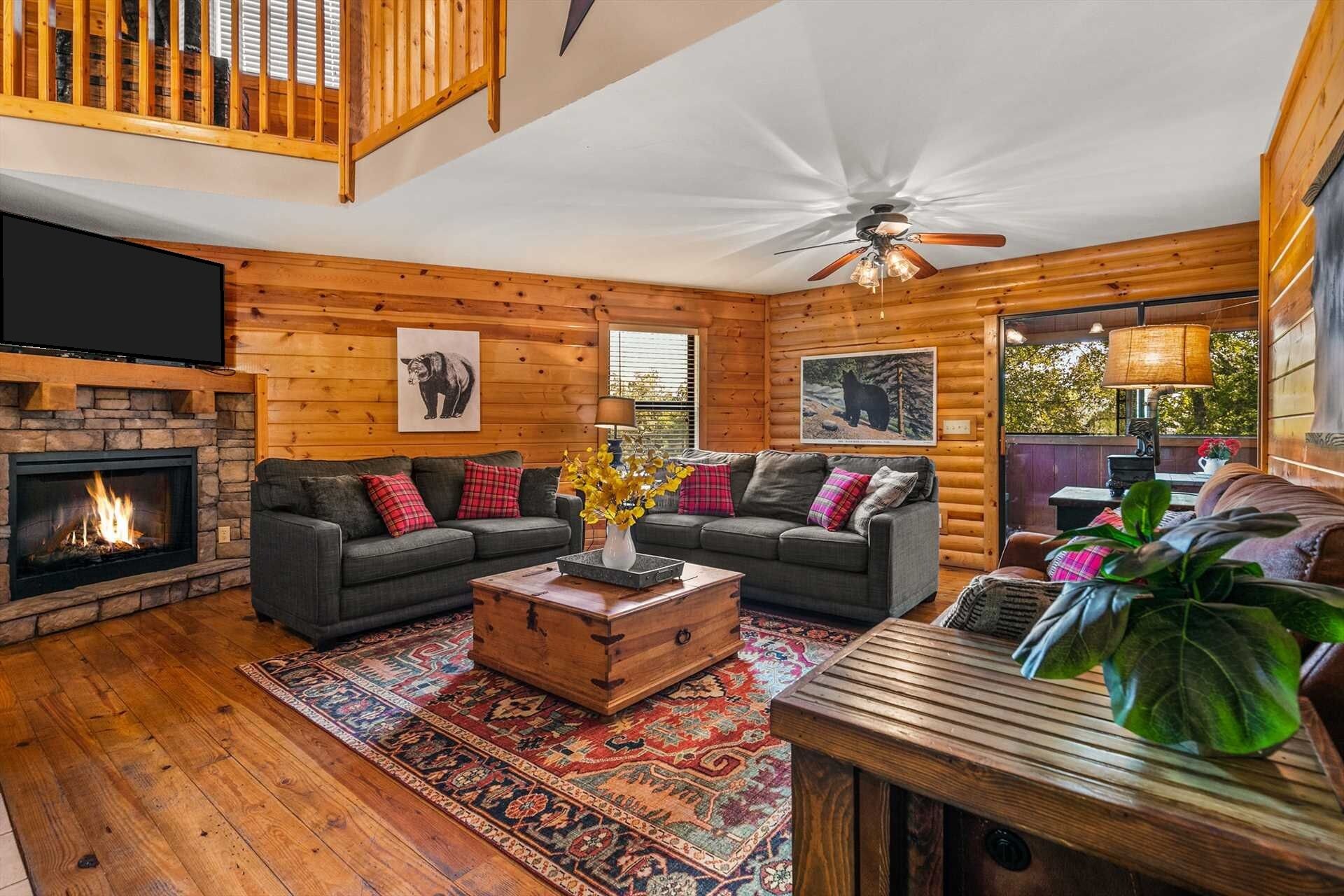 A cozy log cabin interior with a fireplace, two sofas, a coffee table, and a ceiling fan.