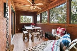 Ein Interieur einer Holzhütte mit einem Esstisch, Bänken, einem Hängesessel und großen Fenstern mit Blick auf Bäume.