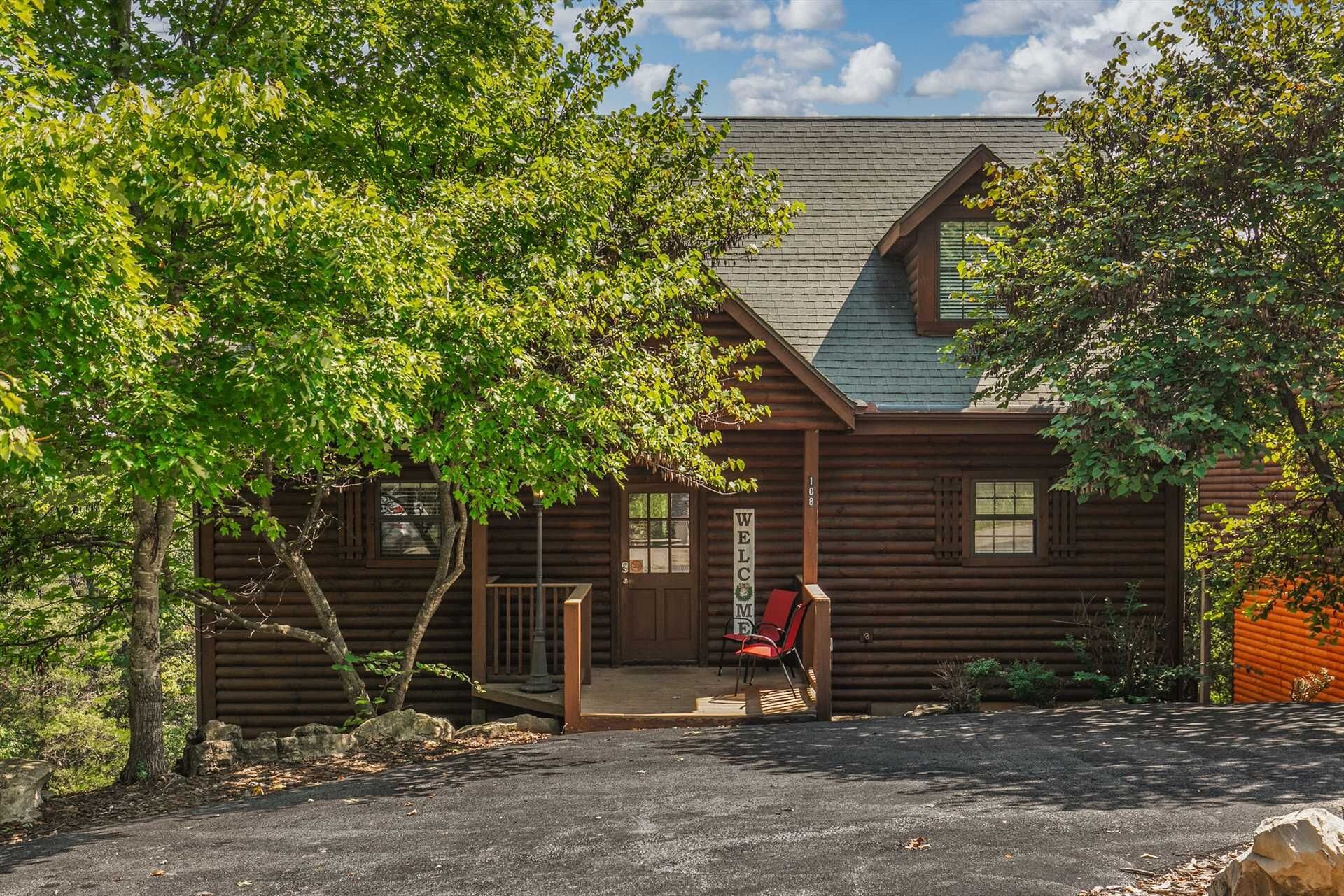 A log cabin with a welcoming sign, a porch with a bench, and a clear sky.