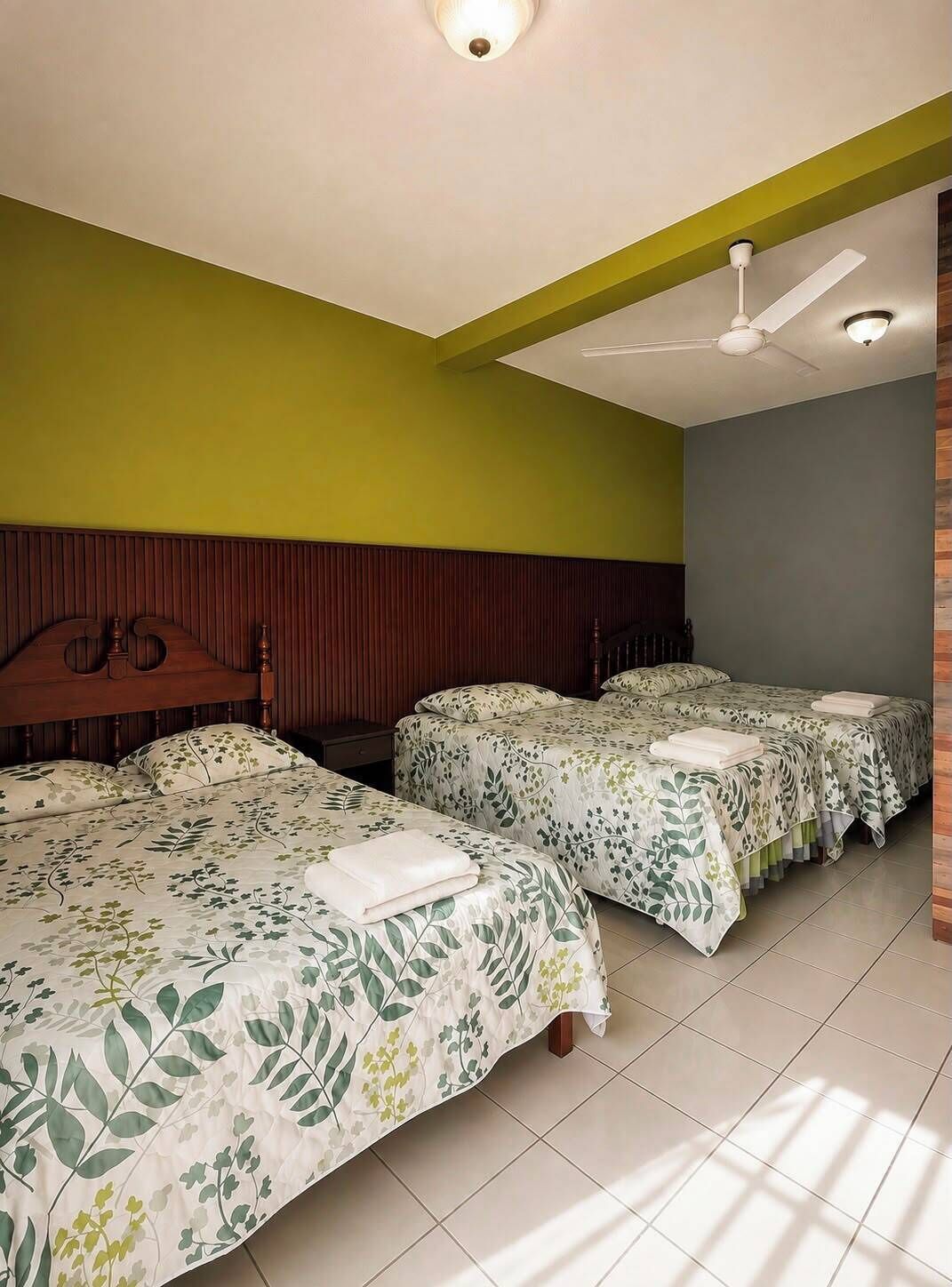 Habitación de hotel con dos camas, cabecero de madera, ventilador de techo y pared verde.