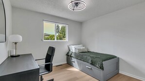 Quarto com cama, escrivaninha, cadeira e janela com vista para árvores.