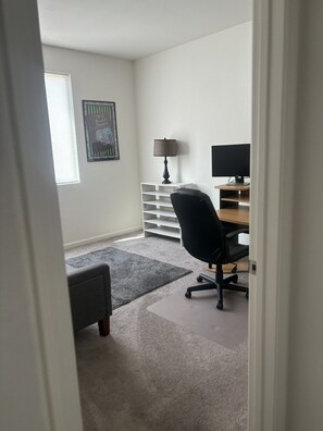 Et værelse med skrivebord, stol, lampe og et billede i ramme på væggen.