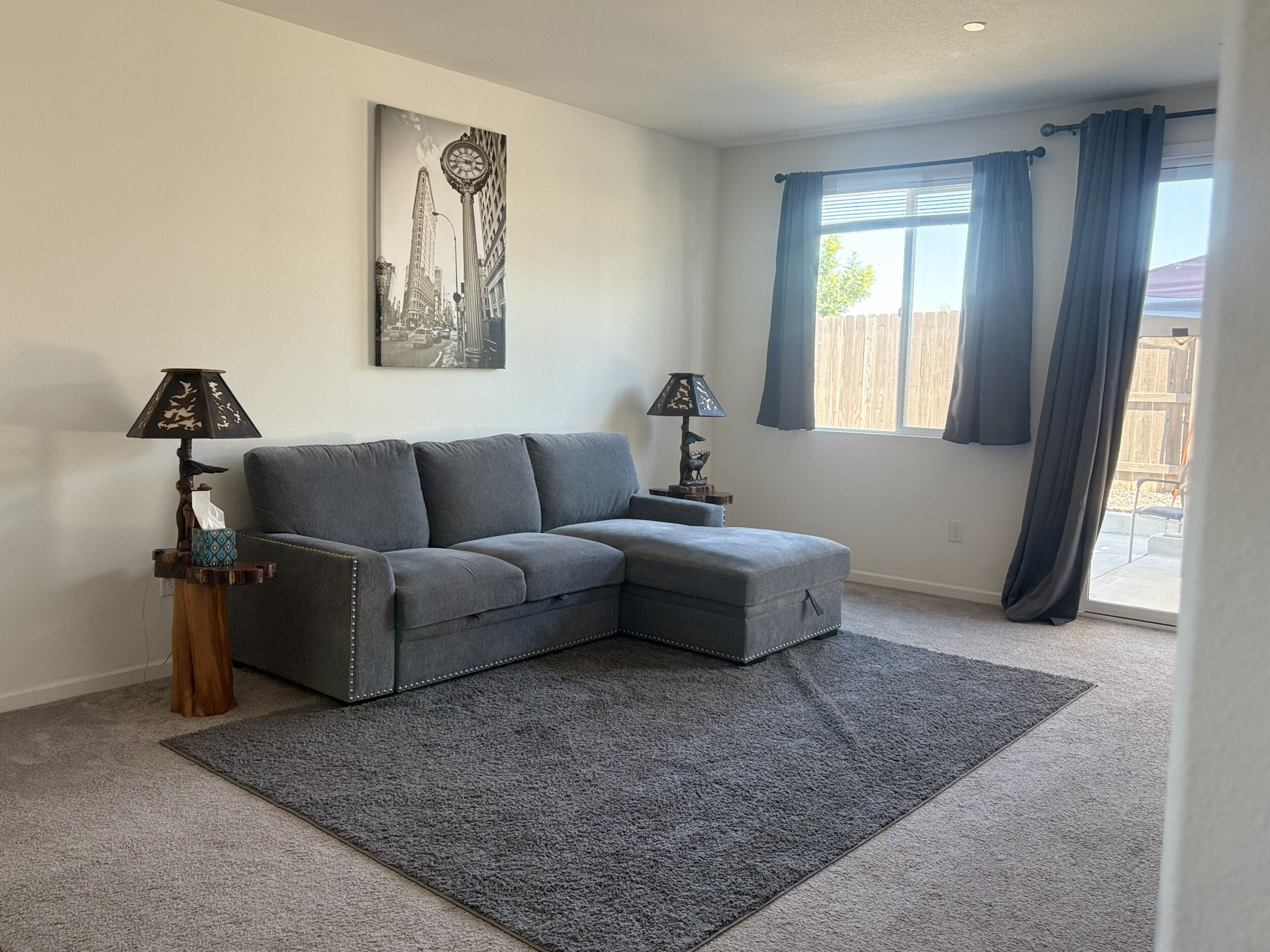 A living room with a grey sectional sofa, two side tables with lamps, a large window with curtains, and a cityscape painting on the wall.