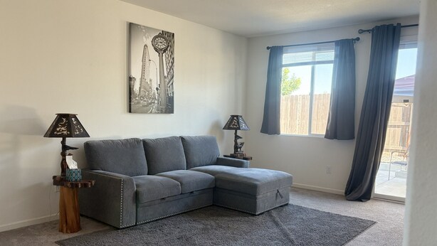 A living room with a grey sectional sofa, two side tables with lamps, a large window with curtains, and a cityscape painting on the wall.