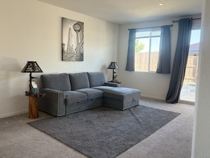 A living room with a grey sectional sofa, two side tables with lamps, a large window with curtains, and a cityscape painting on the wall.