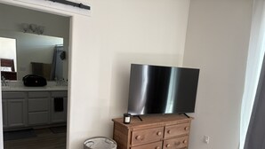 A room with a wooden dresser, a television on top, a grey storage bin, and a bathroom visible through an open door.
