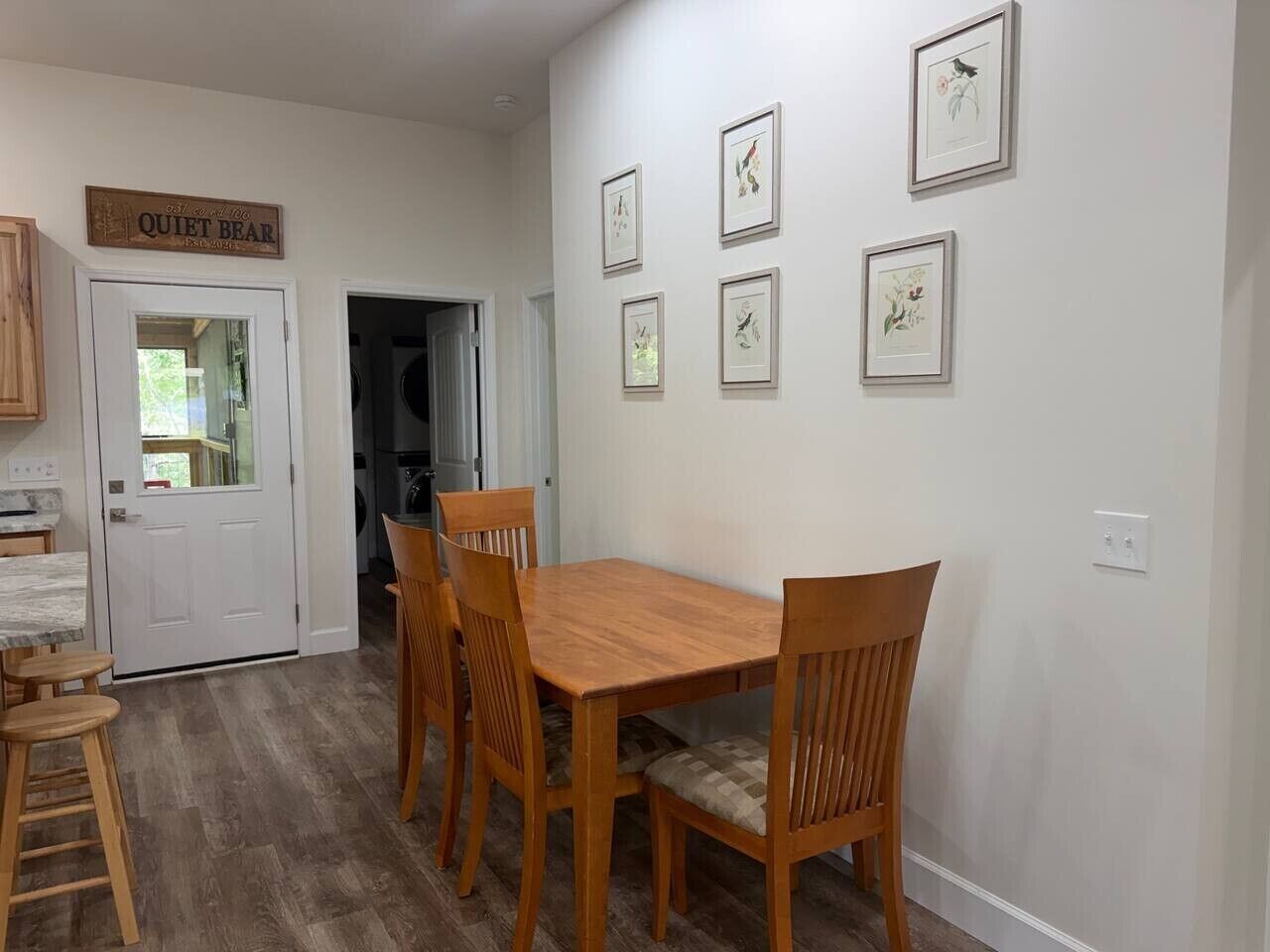 A dining area with a wooden table and chairs, a sign above the door, and framed artwork on the wall.