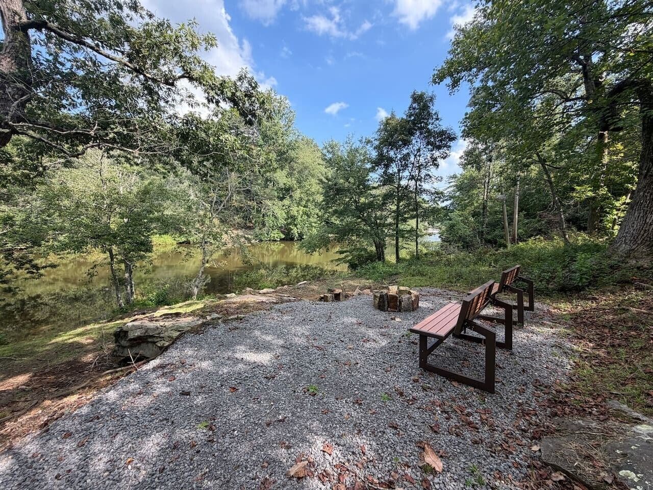 A gravel area with benches and a fire pit, surrounded by trees and a body of water.